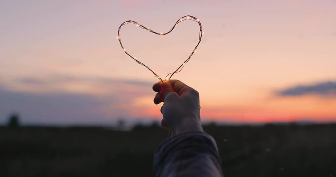 Simbolo del cuore fatto da una striscia di luci led tenuto da una mano di una ragazza di fronte al cielo sfuocato. San Valentino concetto.