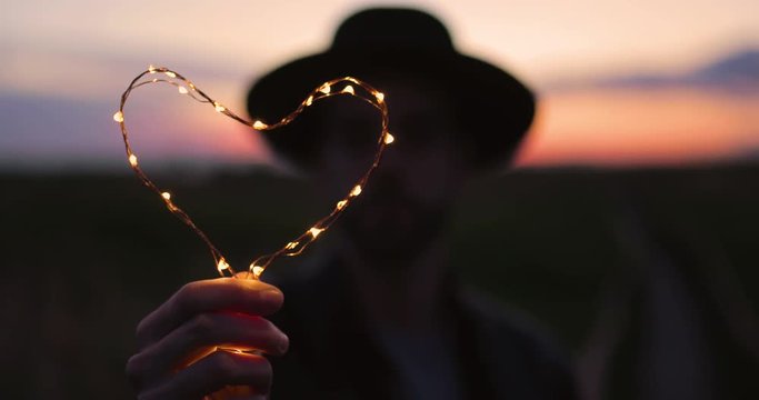Simbolo del cuore con luci led, tenuto in mano da un ragazzo in silhouette con cappello di fronte al tramonto.
