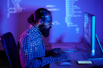 Bearded developer in eyeglasses sitting at the table in front of computer monitor typing on computer keyboard and developing new software