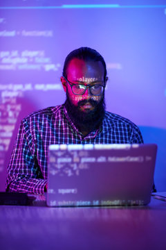 Bearded Programmer In Eyeglasses Concentrating On His Work, He Sitting At The Table Working On Laptop In Dark Office