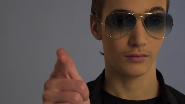 Beautiful teenager pointing finger forward at studio. Handsome boy in dark sunglasses. Teenager in stylish fashionable glasses. Close-up portrait of a young man in beautiful glasses. Guy says yo ...