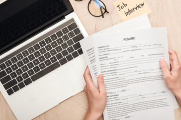 cropped view of recruiter holding resume templates near sticky note with job interview lettering and laptop