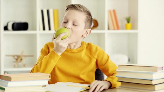 schoolboy taking green and tasty apple and eating while showing thumb up at home 