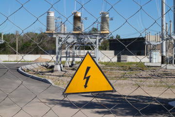 Electrical hazard sign on metal grid. On the background of unfocused high-voltage electrical substation. Concept: danger of electric current, death from electric shock