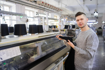 a young man analyzing a knitted piece of cloths near industrial knitted machines with black thread in cones