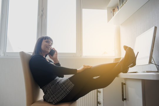 lateral view of a hot brunete talking at the phone in office outfit sitting at the table with a computer and a laptop on it