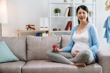 Fototapeta premium Pregnant asian young lady eating fresh fruits living healthy lifestyle taking care of child. elegant woman holding apple face camera smiling hands touch baby in belly sitting on couch sofa at home.