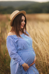 Tender pregnant woman walk in a field. Lady with hat in blue dress. Relax time. Woman is waiting for a baby