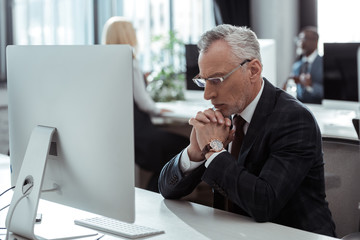 selective focus of pensive mature businessman in glasses sitting near computer monitor and multicultural colleagues in office