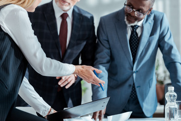 cropped view of businesswoman gesturing near multicultural partners while holding clipboard