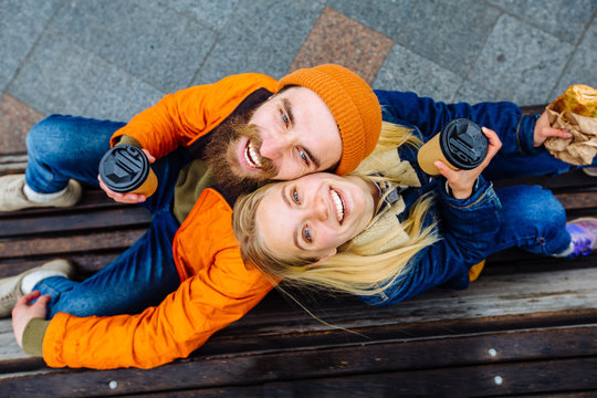 Stylish hipster couple laughing, joking spending time together outdoors. Top view of attractive woman and handsome bearded man are sitting back to back on a bench with coffee cupin the city street