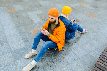 Hipster beard man and blond woman sitting back to back on a longboard Couple using smartphones.Concept of phone addiction. Face absorbed by the phone Stylish and fashionable couple in a urban location