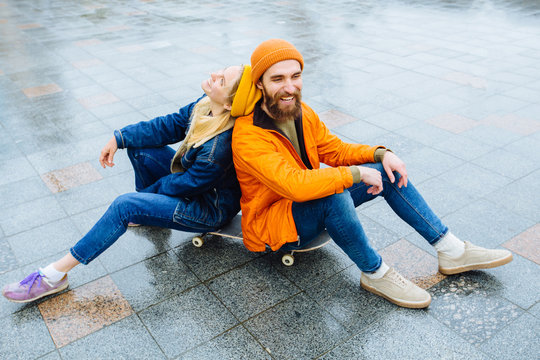 Hipster baerd man and blond woman sitting back to back on a longboard on a concrete surface. Stylish and fashionable couple, shoot in a urban location.