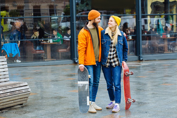 Acrive hipster beard boy and girl friends in bright outwear holding skateboard and longboard looks...