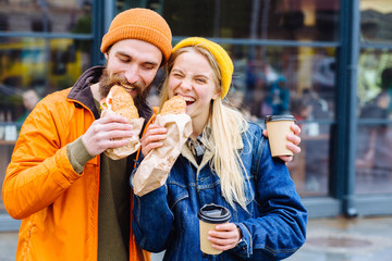 Happy loving couple enjoying breakfast in the city,having fun. street fast food concept. Stylish hipster man and blond woman eating delicious burger, sitting in city street.