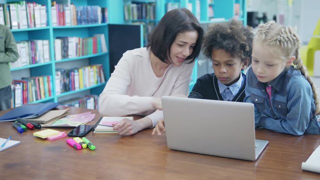 Middle aged female teacher helping two elementary students, mixed race schoolboy and blonde girl, to solve tasks on laptop computer, boy browsing through book in background