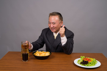 Mature Asian businessman sitting with wooden table against gray 
