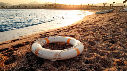 Closeup image of white plastic life saving ring lying on the sandy sea beach against beutiful sunset over the water
