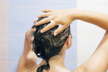 Naklejka premium Young happy woman washing her hair with shampoo, hands with foam closeup. Back of beautiful brunette girl taking shower and enjoying relax time. Body and hair hygiene, lifestyle concept