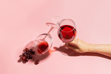 Female hand holding a glass with red wine and bunch of grapes on a gentle pink background....