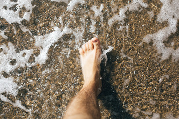 Feet standing in waves foam on sand, top view.  Man foot in warm water waves, relaxing on beach at sea. Summer vacation. Copy space