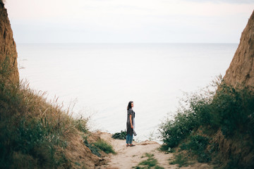 Stylish hipster girl walking to sea between sandy cliffs with grass at sunset. Happy young boho woman exploring tropical island at cliff beach in evening. Summer vacation. Space for text.