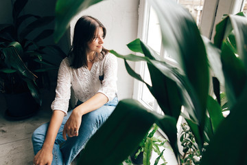 Stylish hipster girl sitting on floor and looking at white rustic window with green plants in modern cafe. Fashionable woman relaxing in soft light at wooden window and green leaves indoors