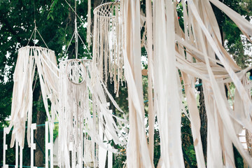 Stylish boho decor on trees. Modern bohemian decoration of white macrame and ribbons, hanging on branches in summer park. Wedding decor and arrangement. Stylish arch for ceremony