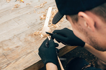 Wooden workshop. Hands carving spoon from wood, working with chisel close up. Process of making wooden spoon, chisel, pencil, compass, ruler on dirty table with shavings