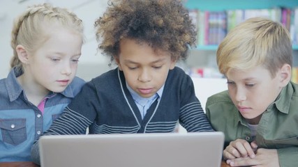 Tracking left medium shot of three curious diverse students studying on laptop computer together sitting at desk in elementary school classroom - Powered by Adobe