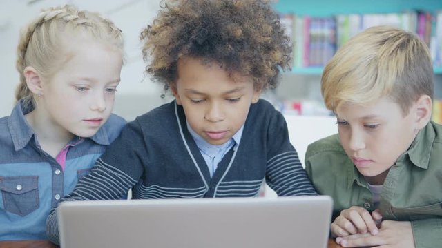 Tracking left medium shot of three curious diverse students studying on laptop computer together sitting at desk in elementary school classroom