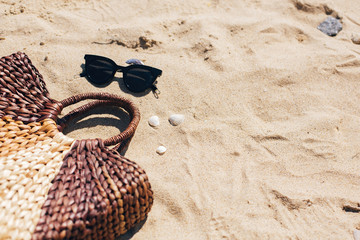 Summer Vacation. Stylish sunglasses and straw bag on sandy beach with sea shells. Girl accessory on...