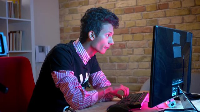 Closeup shoot of young indian attractive male typing on the computer sitting indoors in the cozy apartment with neon light