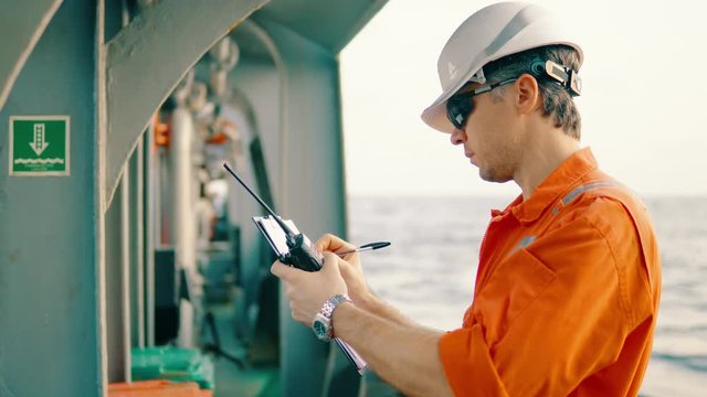 Marine chief officer or chief mate on deck of ship or vessel. He fills up ahts vessel checklist. Ship routine paperwork. He holds VHF walkie-talkie radio in hands.