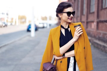 Portrait fashion woman in sunglasses walking on street . She wears yellow jacket, smiling to side.