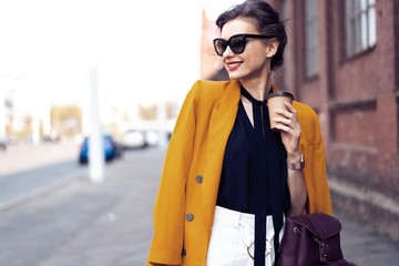 Portrait fashion woman in sunglasses walking on street . She wears yellow jacket, smiling to side.