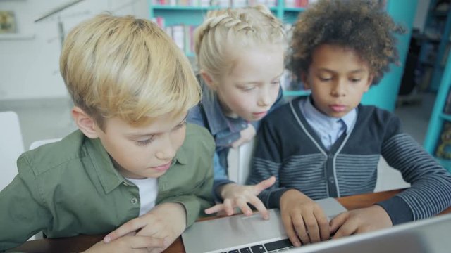 Cheerful schoolgirl helping her male friends to fulfill task or play educational game on laptop computer in elementary school classroom