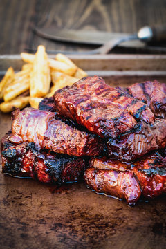 Bbq boneless beef ribs with barbecue sauce and potato wedges over a rustic background. Extreme shallow depth of field with blurred background and selective focus on front of meat