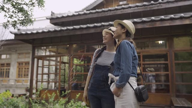 two Asian woman friends tourists stand with hands point at and talk about surroundings while standing by historic wooden house japanese style in kyoto japan. young girls travel visiting monument.