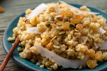 Cooked bulgur with raisins, seeds and coconut in a blue plate on a wooden table. Sweet bulgur porridge close up, Turkish cuisine, dark tones.