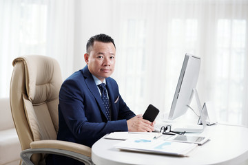 Portrait of Asian office worker sitting on chair at workplace in front of computer with mobile phone and looking at camera at office