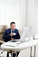 Asian mature businessman sitting at the table looking at computer monitor holding cup and drinking coffee at office