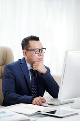 Serious confident businessman wearing eyeglasses working on computer while sitting at desk at office