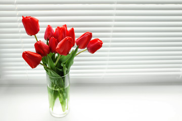 Bouquet of beautiful tulips in vase near window with blinds. Space for text