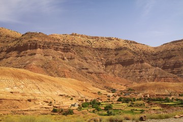 Old berber village oasis with houses build of clay bricks in front of impressive high rugged red mountain face, Gorges du Dades, Morocco