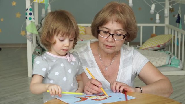 Good looking caucasian grandmother with little blonde granddaughter sit at children's table and paint goldfish on sheet of paper with colored pencils. Children's creativity. Close-up.