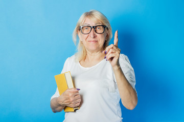 Old woman with glasses holds a book in her hands and shows a hand gesture on a blue background. Concept Knowledge and power of reading and education