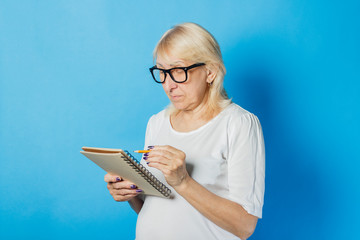 Old lady with glasses holds a notebook and pen in her hands against a blue background. Concept note taking, crossword, sudoku, stacking list, bad memory