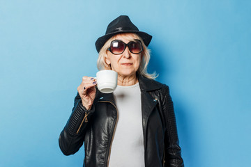 Old lady with glasses, a leather jacket and a hat is holding a cup of coffee against a blue...