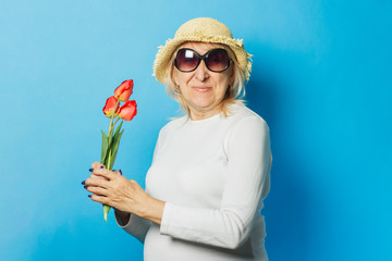 Old woman in a straw hat and sunglasses is holding a bouquet of tulips on a blue background. Concept of the onset of spring, summer time, vacation, camping